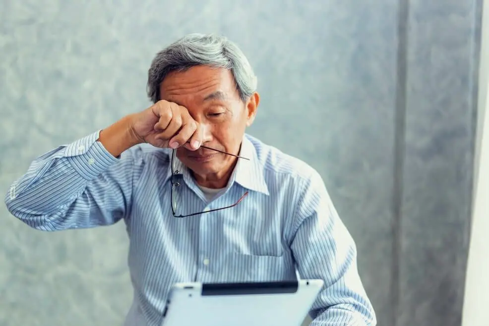 Elderly man feeling tired or stressed, rubbing his eyes while holding glasses and paperwork, possibly needing a visit to the Premier Eye Clinic.